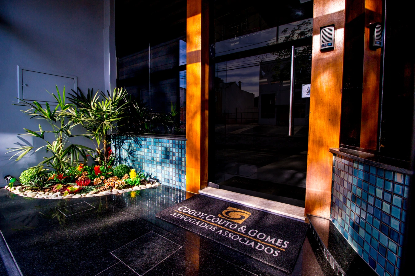 Entrada principal do escritório Cogoy, Couto & Gomes, com fachada de vidro e tapete personalizado com o logotipo.
