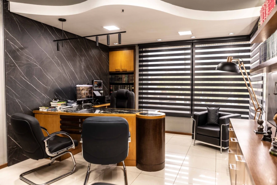 Vista geral do escritório de Carlos Cogoy com estante de livros, persianas modernas e mesa de atendimento em madeira com acabamento em mármore.
