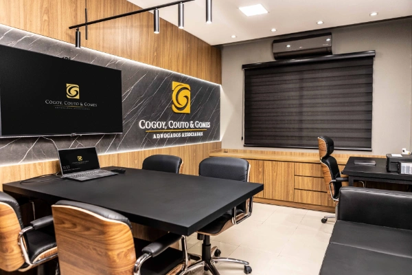 Sala de reuniões do CCG com logotipo na parede, mesa retangular preta e cadeiras executivas com encosto de madeira.